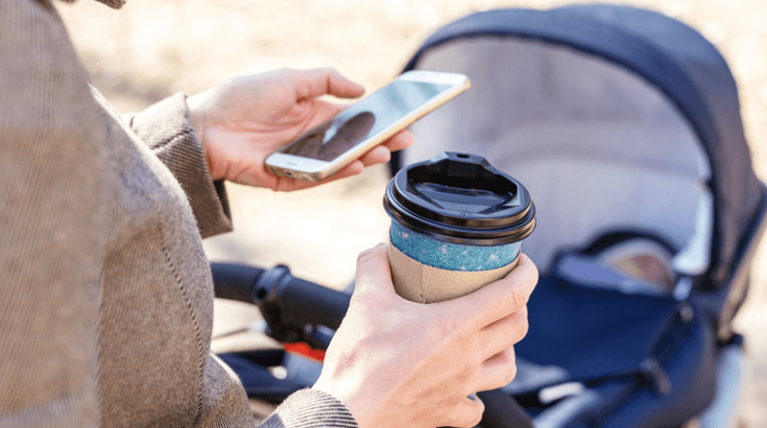 A woman pushes a pram while holding her mobile phone and a takeaway coffee cup.