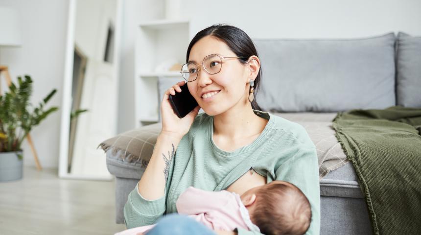 Mother breastfeeds baby while talking on the phone and smiling in her living room.