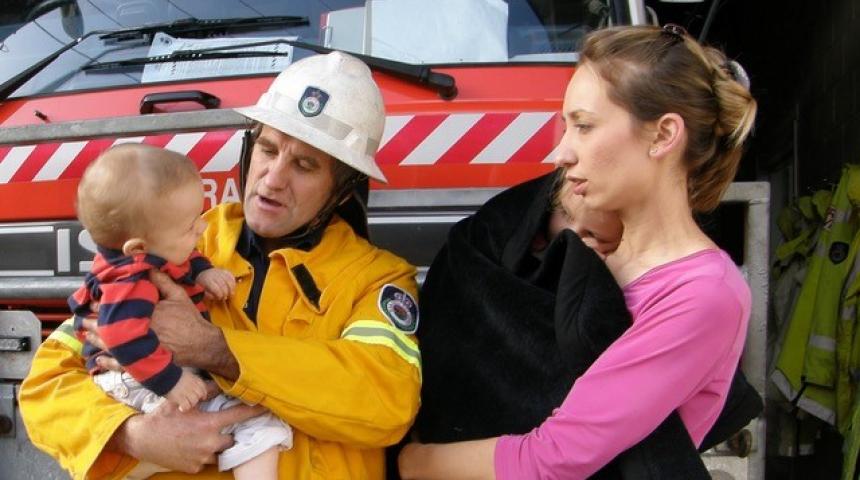 A mother and fireman hold two young children and stand in front of a fire truck.