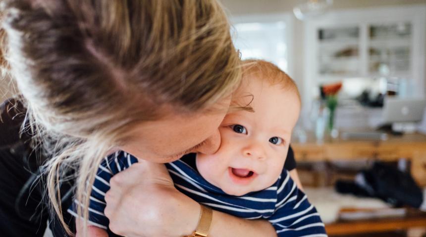 A mid-range photograph of a mother holding her smiling baby and kissing his cheek.