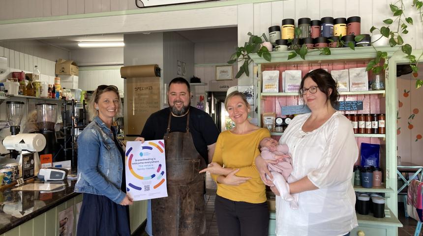 Four people, one holding a baby, standing in a cafe and smiling at the camera.