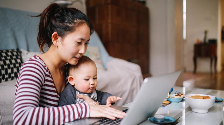 A mother holds her baby and uses her laptop