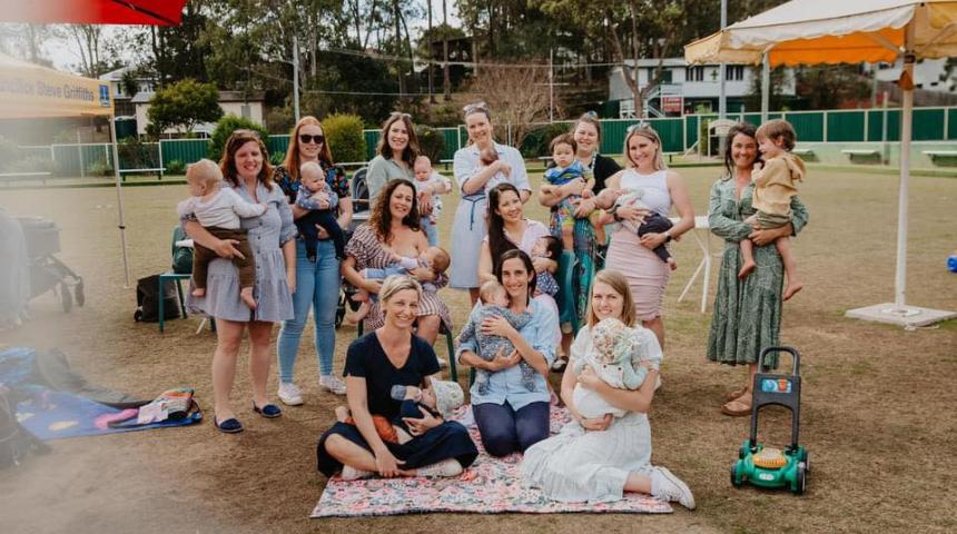 Group of mums and babies/children facing the camera enjoying time together. There are picnic mats and toys dispersed around. 