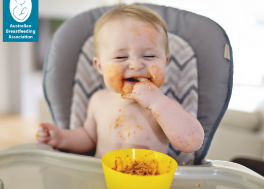Baby in a high chair grinning with finger in mouth and orange food around their mouth.