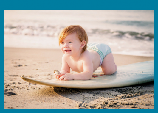Toddler looking cheerful, lying on a surfboard on the sand with the ocean behind them