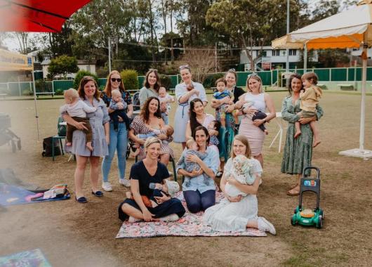 Group of mums and babies/children facing the camera enjoying time together. There are picnic mats and toys dispersed around. 