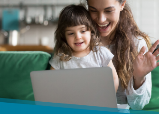 A mother waves at a laptop screen while her toddler sits on her lap