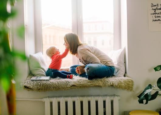 Mother and toddler playing together while sat in a window seat. Toddler is touching mum's face playfully..
