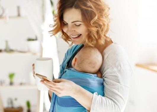 Photo of a mum wearing a baby in a sling, looking at her phone and holding a coffee mug