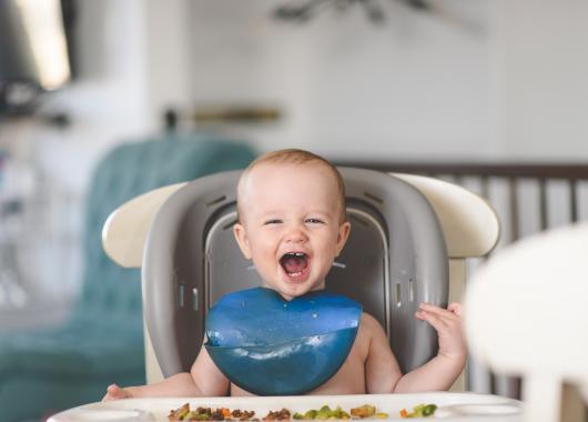 Baby sitting upright in high chair mouth open and smiling