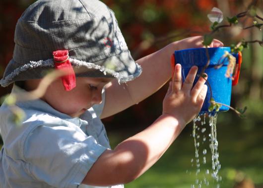A toddler wearing a green bucket hat and a blue t shirt. He is holding up a small blue bucker with small sticks and leaves sticking out, while water flows out of holes in the bottom of the bucket.