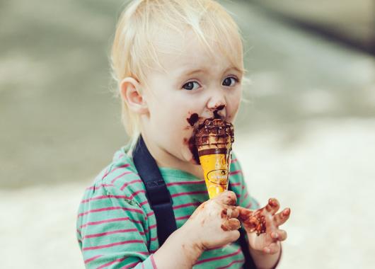 A cute blonde toddler eats a chocolate ice cream in a cone. The ice cream is smeared around his mouth and across his hands.