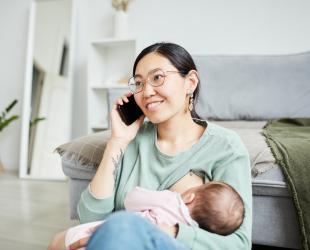 Mother breastfeeds baby while talking on the phone and smiling in her living room.