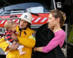 A mother and fireman hold two young children and stand in front of a fire truck.