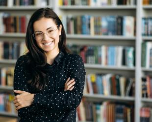 Portrait of young woman smiling at the camera with bookshelves behind. nding in modern office or university library with arms crossed, show self confidence