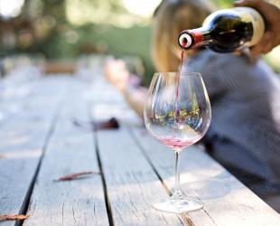 Red wine being poured from a bottle into a glass. The glass is on an outdoor table with people in the background at a party or celebration.