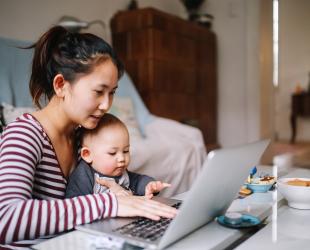 A mother holds her baby and uses her laptop