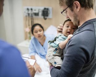 Father holding young baby with mother in background in a hospital bed as she awaits a colonoscopy