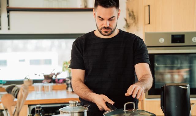 An image of a man in a black tshirt in the kitchen. He is cooking at the stove with pots and pans.