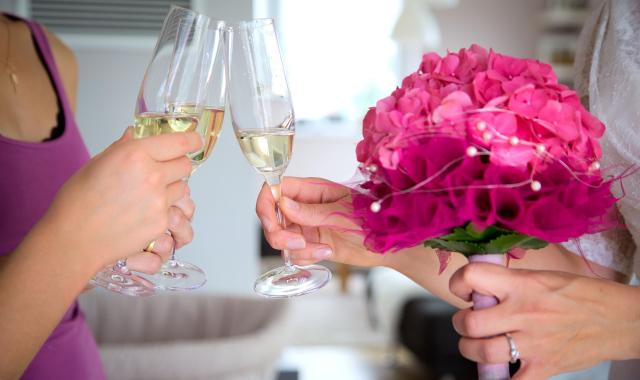 Image of women clinking champagne flutes. One woman is a bride in a white dress holding a bouquet of pink flowers.