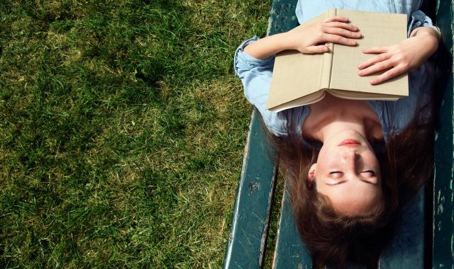 A picture of a woman lying on a bench in a grassy space. We are looking at her from above. Her eyes are peacefully closed and she's clasping a book to her chest.