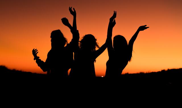 An image of 3 women dancing with their hands freely moving in the air. They are silhouetted against an orange sunset.