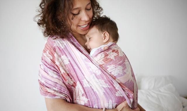 A woman smiles down at the young baby she holds in a pink floral baby carrier.