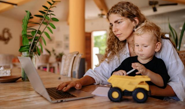 mum sitting at table one hand typing on laptop with a 1-year sitting on her laptop holding a toy truck