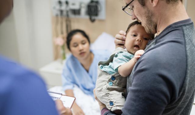 Father holding young baby with mother in background in a hospital bed as she awaits a colonoscopy