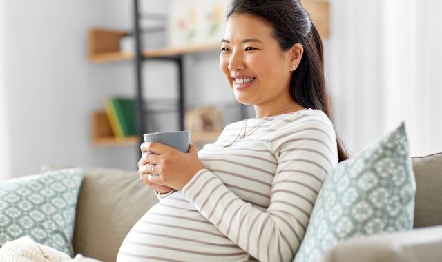 A pregnant woman sits on the couch with a cuppa and smiles