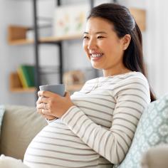 A pregnant woman sits on the couch with a cuppa and smiles